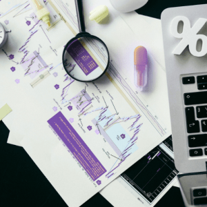 Análise de gráficos e dados de mercado em uma mesa com computador, gráficos, caneta roxa, lente de aumento e papel com diversas informações e números